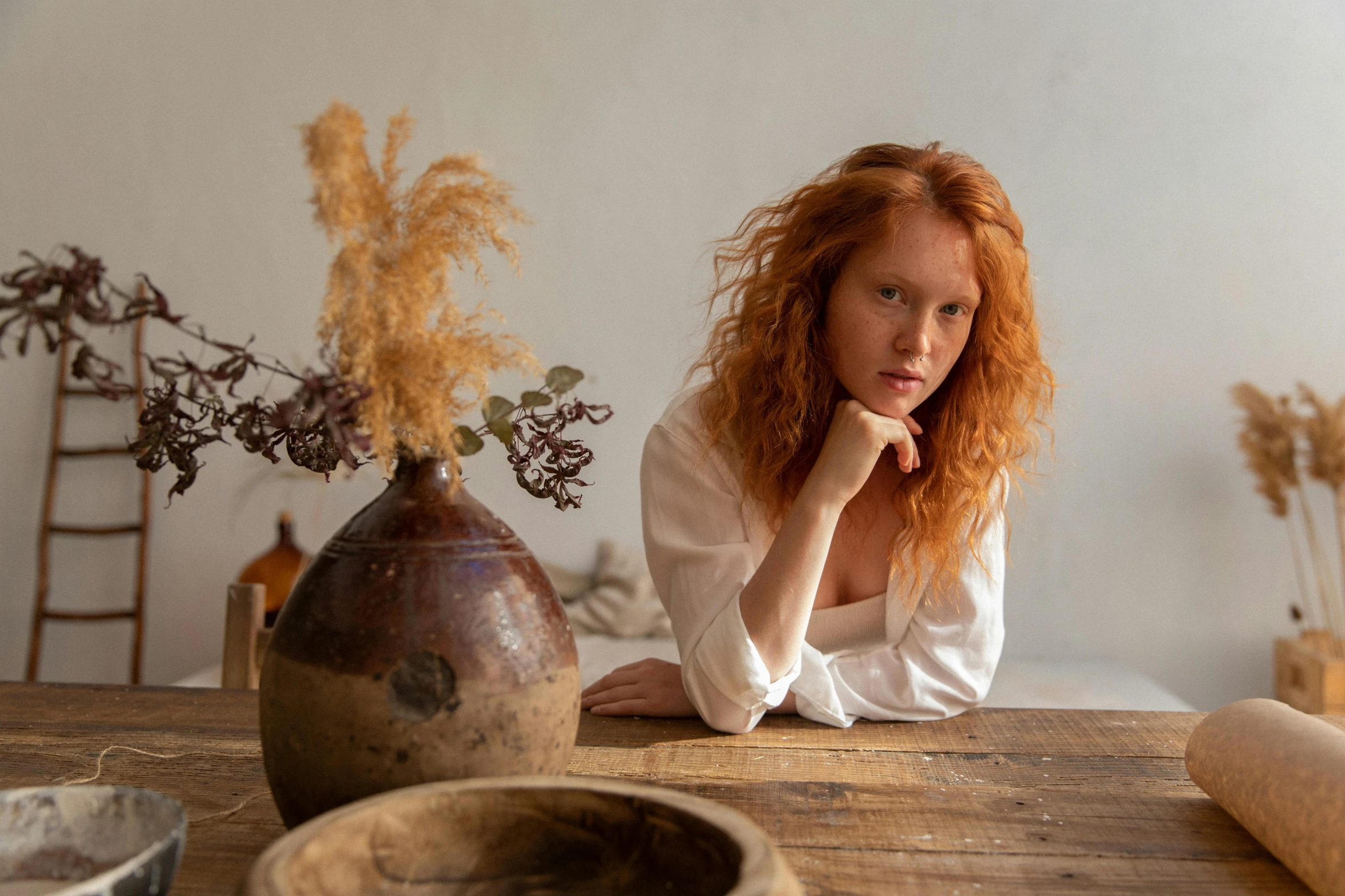 a woman sitting at a table with a vase in front of her, inspired by William Stott, pexels contest winner, renaissance, ginger wavy hair, wearing a linen shirt, photoshoot for skincare brand, natural dull colours