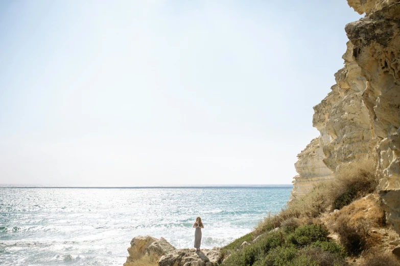 a man standing on top of a cliff next to the ocean, cyprus, conde nast traveler photo, woman on the beach, grey