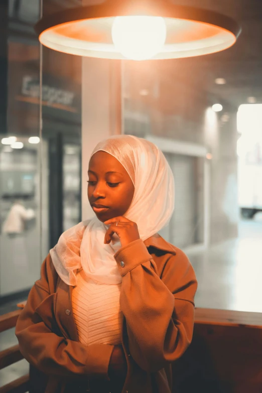 a woman wearing a hijab standing in front of a window, an album cover, pexels contest winner, ( brown skin ), at a mall, thoughtful pose, soft luminescent glow