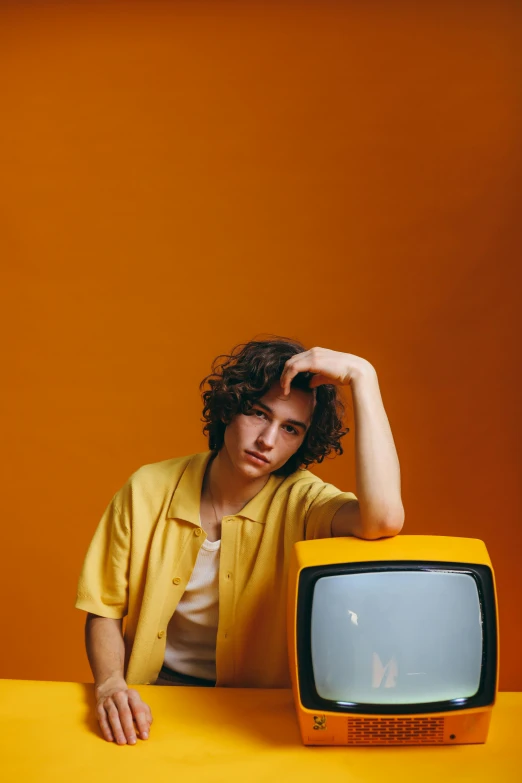 a man sitting at a table with a television in front of him, an album cover, pexels, wavy hair yellow theme, teenage boy, ansel ], asher duran