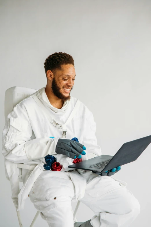 a man sitting in a chair with a laptop, inspired by Dr. Atl, pexels contest winner, afrofuturism, with a lab coat, nasa eva suit, dirty linen robes, looking happy