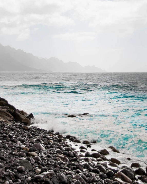 a man standing on top of a rocky beach next to the ocean, azure waves of water, shades of blue and grey, malika favre, a photo of the ocean