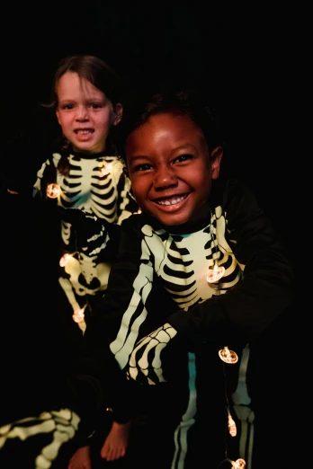 a couple of kids standing next to each other, an album cover, pexels, glowing bones, diverse costumes, both smiling for the camera, dark. no text