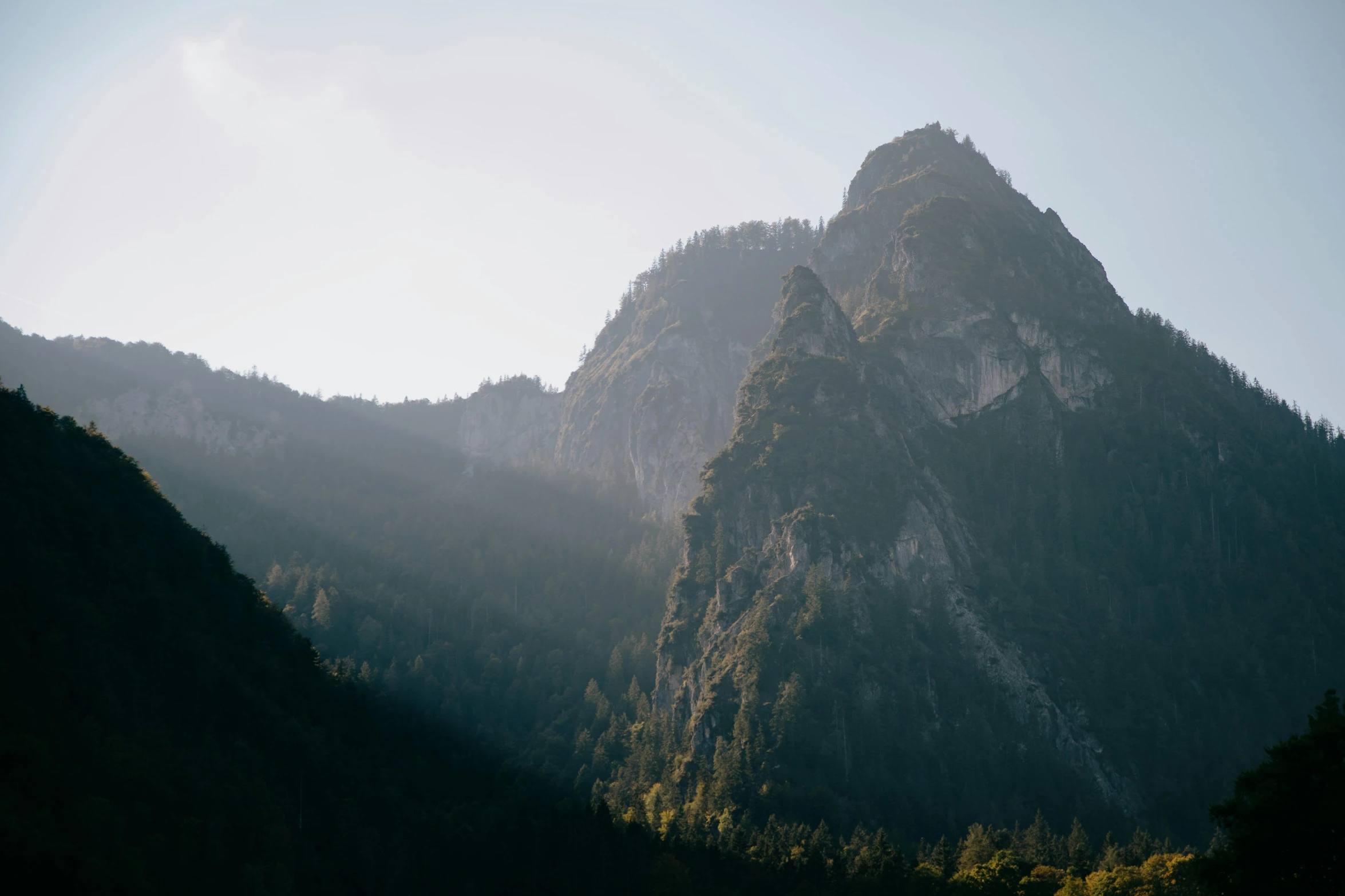 a view of a mountain range with trees in the foreground, unsplash contest winner, very long spires, strong sunlight, minimal shading, steep cliffs