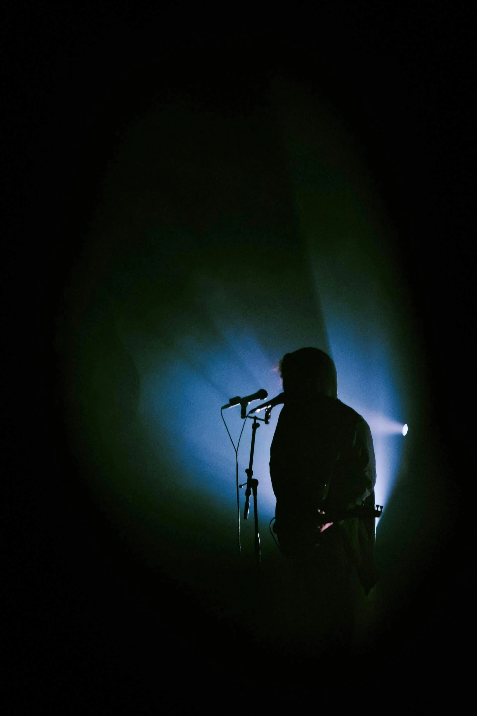 a man standing in front of a microphone in the dark, an album cover, unsplash, crystal castles, concert light, human silhouette, guitarist