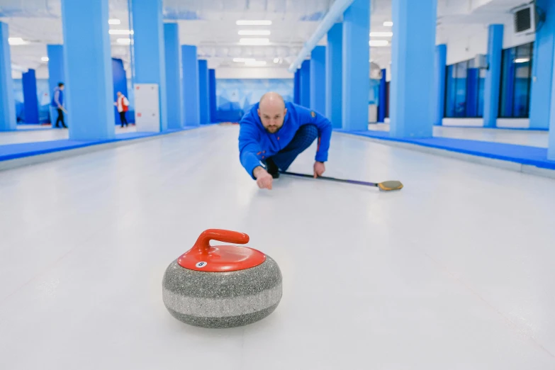 a man bending over to pick up a curling stone, by karlkka, soft surfaces, blue fireball, spacious, looking towards camera