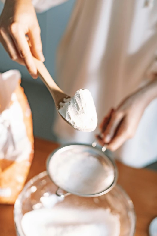 a person holding a spoon over a bowl of food, covered in white flour, detailed product image, skincare, baking a cake