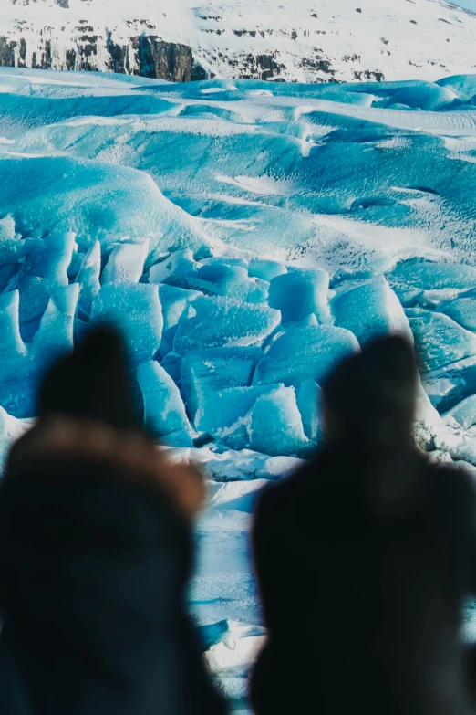 a group of people standing in front of a glacier, rich blue colors, close-up shot from behind, two still figures facing camera, unsplash photography