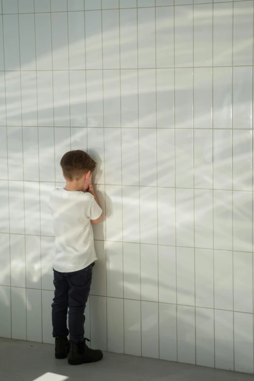 a little boy that is leaning against a wall, inspired by Leandro Erlich, unsplash, interactive art, made of all white ceramic tiles, square lines, facepalm, natural soft light