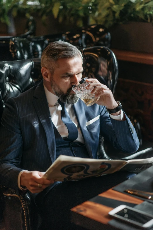 a man sitting in a chair drinking from a glass, a portrait, by Adam Marczyński, pexels, luxurious suit, reading a newspaper, bearded man, beer