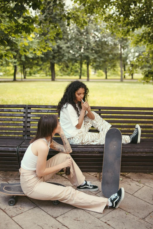 a couple of people sitting on a bench with a skateboard, trending on pexels, lesbian, infp young woman, smoking, park