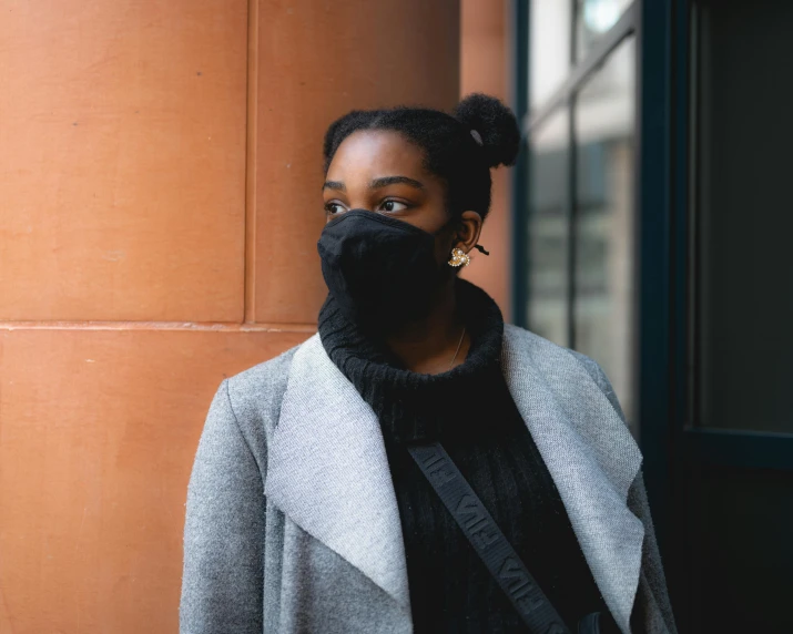 a woman wearing a face mask standing in front of a building, by Nina Hamnett, pexels contest winner, grey turtleneck coat, black girl, over the shoulder, 1 5 0 4