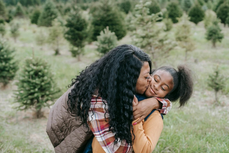 two women hugging each other in a field, by Everett Warner, pexels contest winner, with a kid, black young woman, pine trees in the background, holiday season