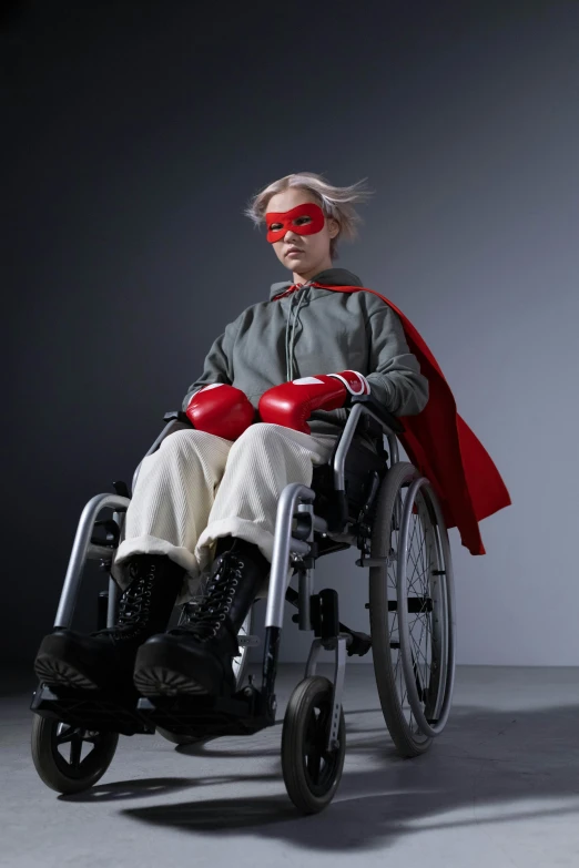 a woman in a red cape sitting in a wheelchair, an album cover, inspired by Jens Ferdinand Willumsen, trending on pexels, japanese dc live-action movie, wearing goggles, dramatic press photo, battle ready