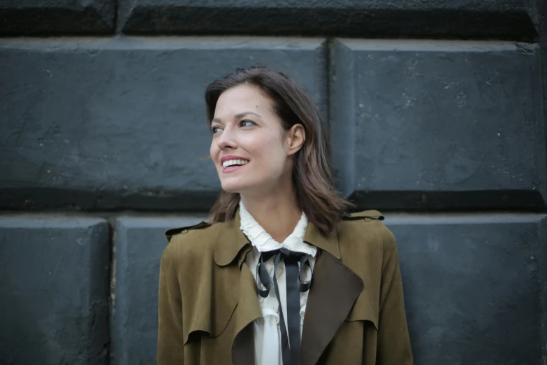 a woman standing in front of a black wall, by Nina Hamnett, pexels contest winner, light brown trenchcoat, smiling slightly, ribbon, brown haired