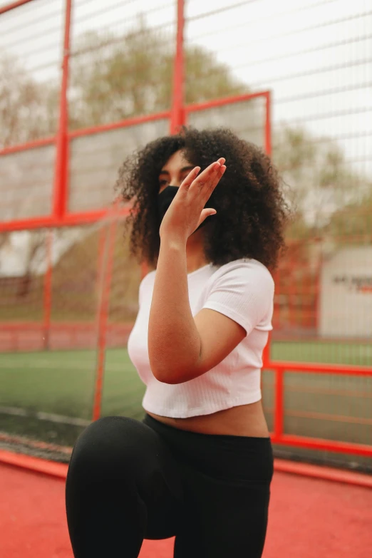 a woman standing on top of a tennis court holding a racquet, an album cover, by Attila Meszlenyi, pexels contest winner, long afro hair, hand over mouth, on a football field, woman in streetwear