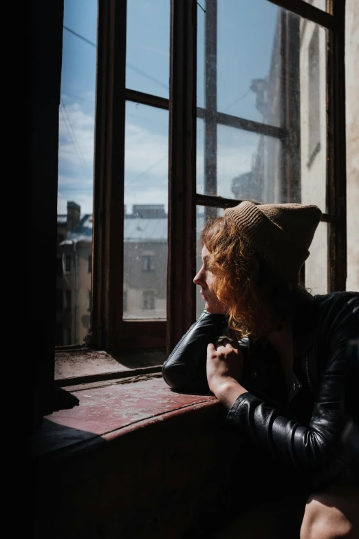 a man sitting on a window sill looking out a window, inspired by Nan Goldin, trending on pexels, a redheaded young woman, wearing a french beret, pale skin curly blond hair, wearing a dark hood