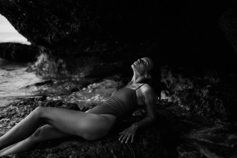 a woman laying on top of a rock next to the ocean, a black and white photo, inspired by Max Dupain, unsplash, figuration libre, in dark cave, athletic fashion photography, bathed in the the glow, model posing