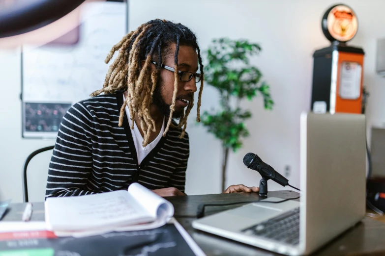 a man sitting at a desk in front of a laptop computer, by Everett Warner, trending on unsplash, figuration libre, rapping into microphone, dreadlock black hair, ai researcher, in a classroom