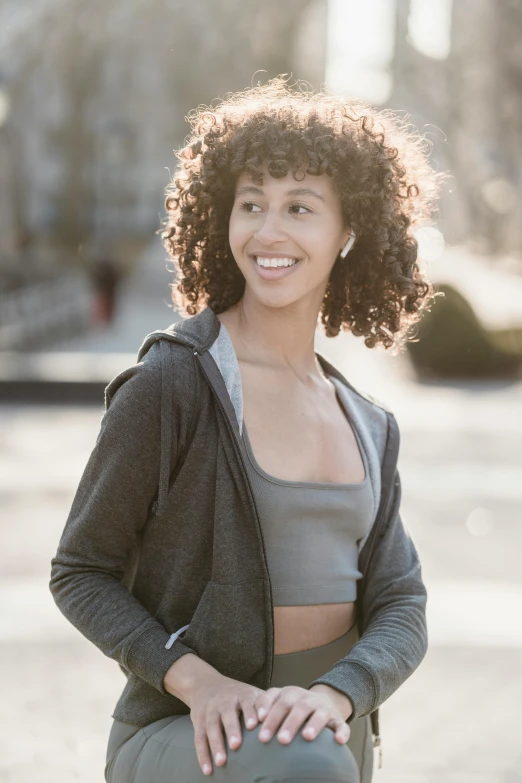 a beautiful young woman sitting on top of a skateboard, by Nina Hamnett, trending on pexels, happening, curly middle part haircut, sports bra, grey jacket, smiling and dancing