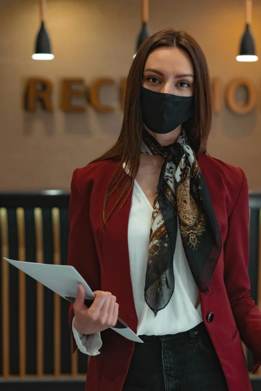 a woman wearing a mask and holding a piece of paper, in a hotel hallway, red scarf, wearing a black blazer, brown
