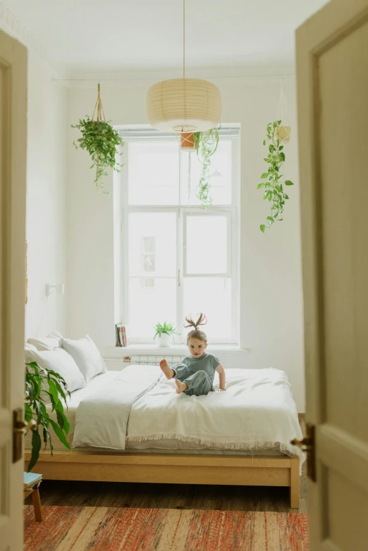 a small child sitting on a bed in a bedroom, pexels contest winner, minimalism, lush plant growth, things hanging from ceiling, city apartment cozy calm, springtime morning