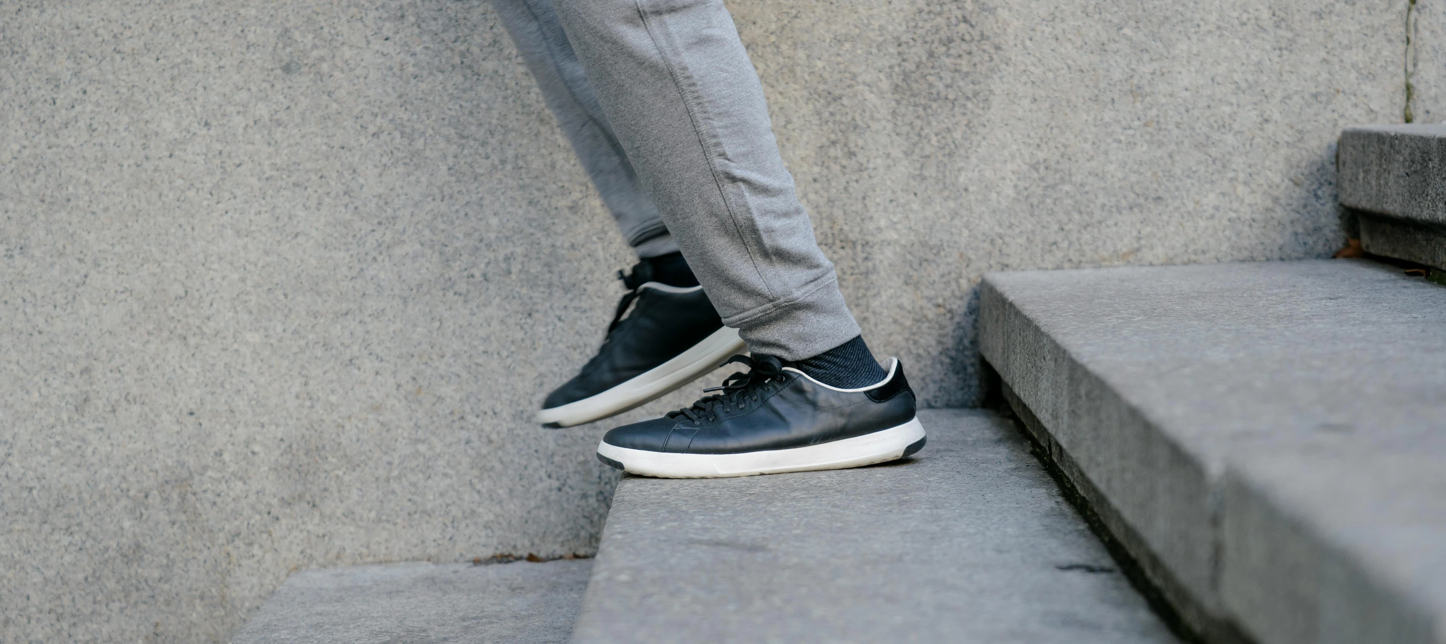 a man riding a skateboard up the side of a cement wall, a black and white photo, grey pants and black dress shoes, focus on sneakers only, standing on rocky ground, wearing leather