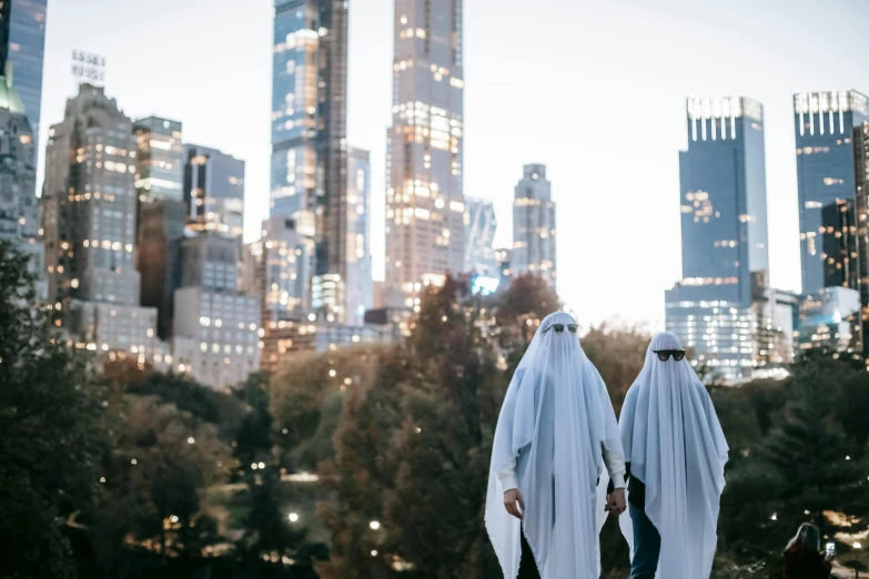 a couple of people that are standing in the grass, inspired by Christo, pexels contest winner, surrealism, halloween ghost under a sheet, new york city as backdrop, white hijab, city buildings on top of trees