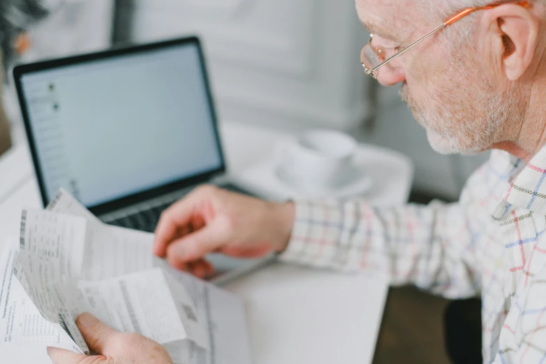 a man sitting at a desk with a laptop and papers, pexels contest winner, two old people, old gigachad with grey beard, lachlan bailey, looking from shoulder