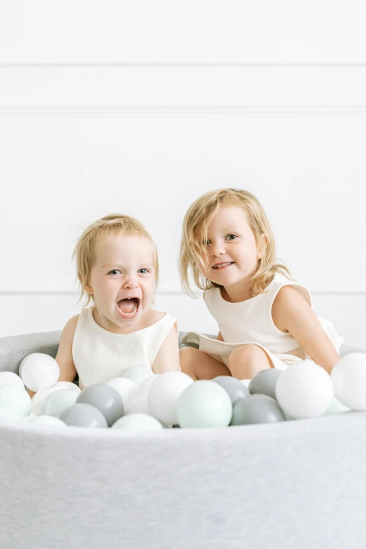 two little girls are sitting in a ball pit, by Eden Box, made in tones of white and grey, mint higlights, high quality product photo, half image