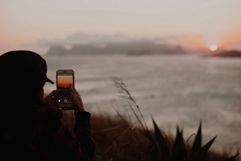 a woman taking a picture of the sunset with her cell phone, a picture, unsplash contest winner, on the coast, slight overcast, looking out at a red ocean, looking at his phone