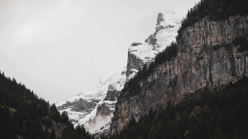 a mountain covered in snow next to a forest, by Daniel Seghers, pexels contest winner, overcast gray skies, cliff side, three fourths view, 4 k hd wallpapear