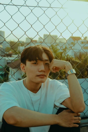 a young man sitting in front of a chain link fence, an album cover, inspired by jeonseok lee, trending on pexels, thick jawline, ( golden hour ), lovingly looking at camera, profile image
