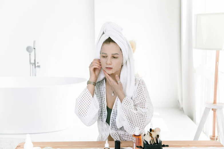 a woman putting makeup on her face in front of a mirror, trending on pexels, process art, wearing a towel, on a white table, manuka, wearing long white robe