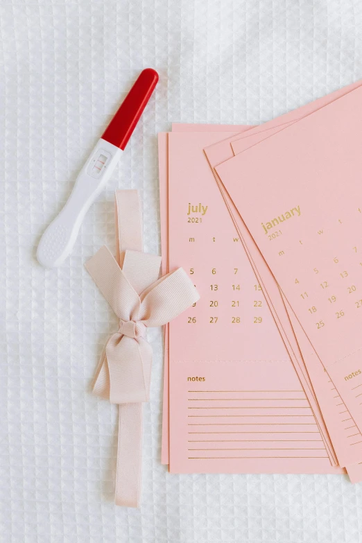 a pink notebook sitting on top of a bed next to a red pen, gold foil, female calendar, ribbon, 3 - piece