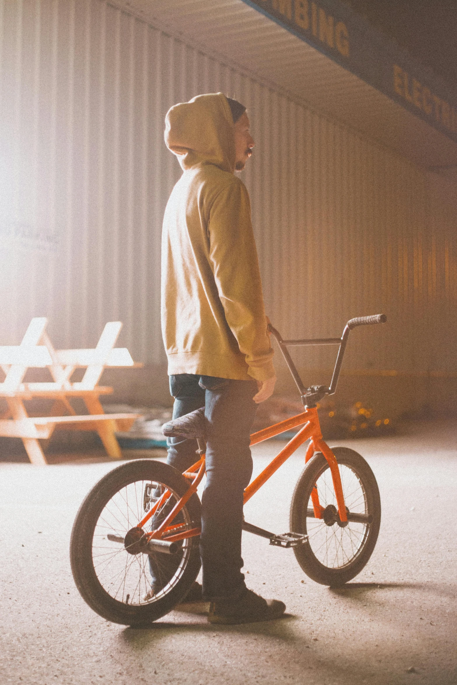 a man standing next to a bike in front of a building, orange hoodie, at a skate park, profile image, hazy