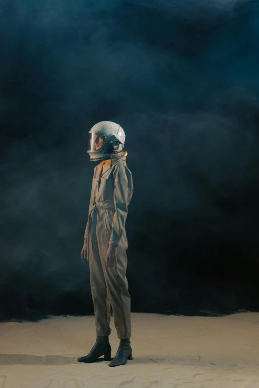 a man standing in the sand with a helmet on, an album cover, by Adam Marczyński, pexels contest winner, sitting sad in spaceship, flight suit, concert, dark backdrop