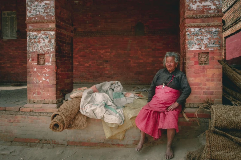 a woman sitting on a bench in front of a brick building, a portrait, pexels contest winner, hyperrealism, nepali architecture buildings, red robes, medium format. soft light, two aboriginal elders