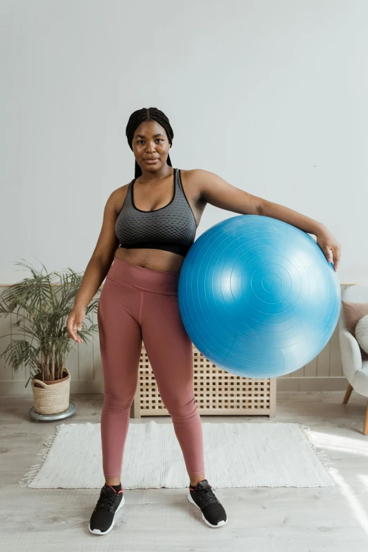 a woman standing in a living room holding an exercise ball, by Matija Jama, pexels contest winner, real life size, african sybil, product introduction photo, half body