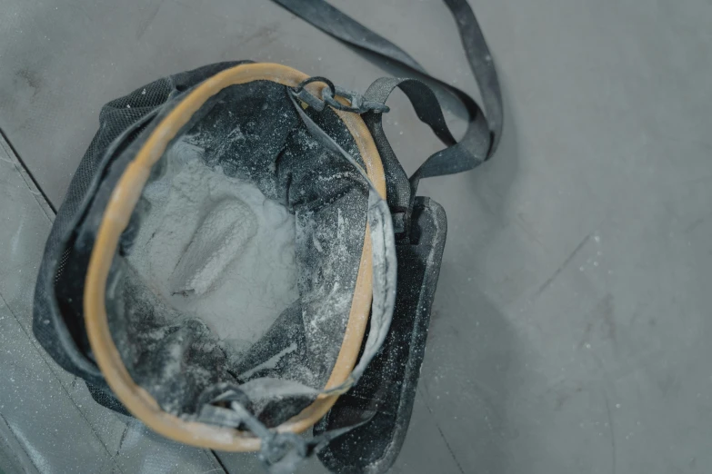 a black bag sitting on top of a cement floor, by Emma Andijewska, plasticien, dust mask, background image, smashed glass helmet, in a workshop