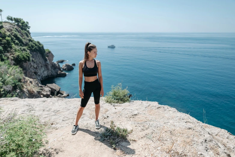 a woman standing on a cliff overlooking the ocean, by Emma Andijewska, pexels contest winner, happening, wearing fitness gear, avatar image, wearing black shorts, full body photo