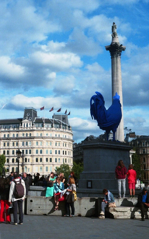 a group of people standing around a statue, blue phoenix bird, london streets in background, square, rooster