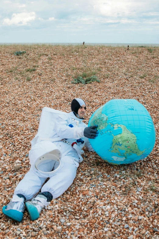 a man laying on top of a beach next to a blue ball, an album cover, inspired by Scarlett Hooft Graafland, unsplash, fantastic realism, beautiful woman in spacesuit, the thames is dry, wearing flight suit, approaching earth