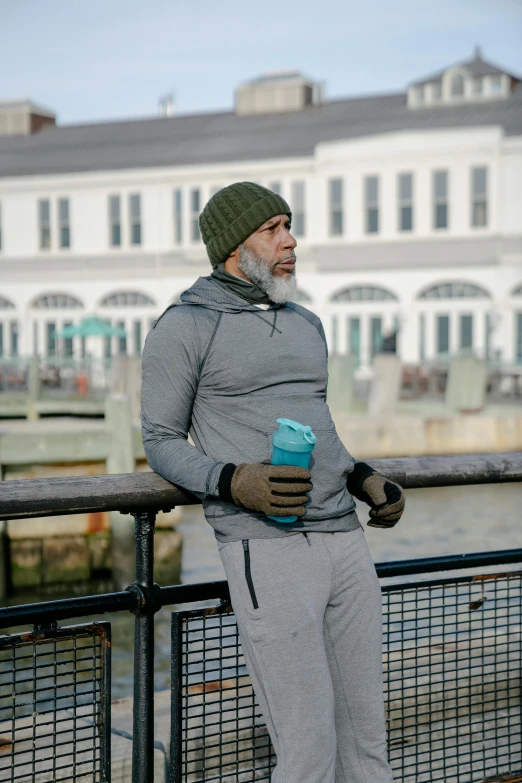 a man standing on a bridge next to a body of water, wearing teal beanie, holding a drink, gray beard, wearing fitness gear