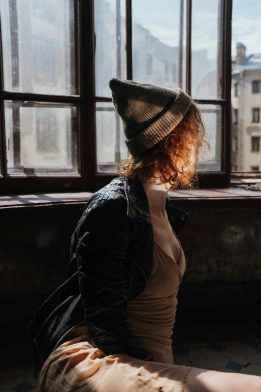 a woman sitting on the floor in front of a window, inspired by Elsa Bleda, trending on pexels, renaissance, beanie, ginger wavy hair, standing in abandoned building, profile pic