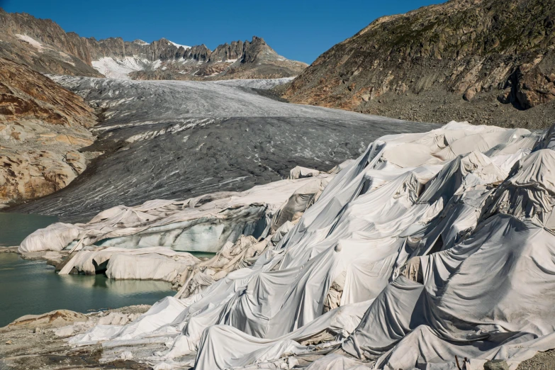 a mountain covered in tarp next to a body of water, by Alison Geissler, unsplash contest winner, land art, glaciers, marble sculptures, the pits of hell, lined in cotton