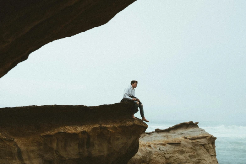 a man sitting on top of a rock next to the ocean, by Ryan Pancoast, pexels contest winner, large overhangs, transparent background, liminal space aesthetic, natural cave wall