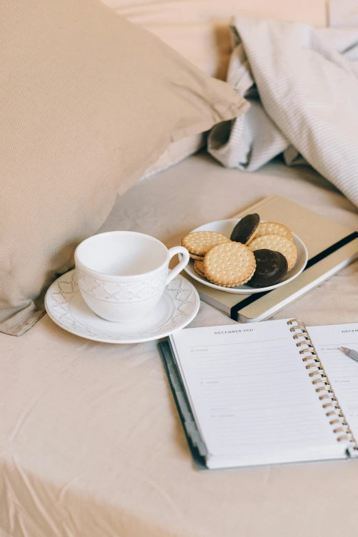 a laptop computer sitting on top of a bed next to a cup of coffee, by Nicolette Macnamara, pexels contest winner, light beige pillows, snacks, luxury journal cover, tea cup