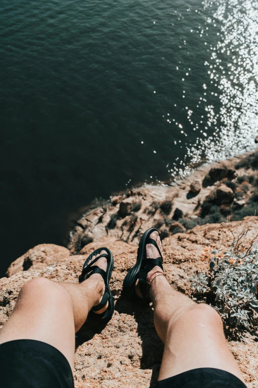 a person sitting on top of a rock next to a body of water, slippers, top down shot, view(full body + zoomed out), steep cliffs