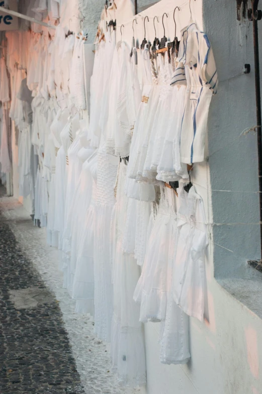 a row of wedding dresses hanging on a wall, inspired by Modest Urgell, renaissance, streets of salvador, white color, dusk setting, the western wall